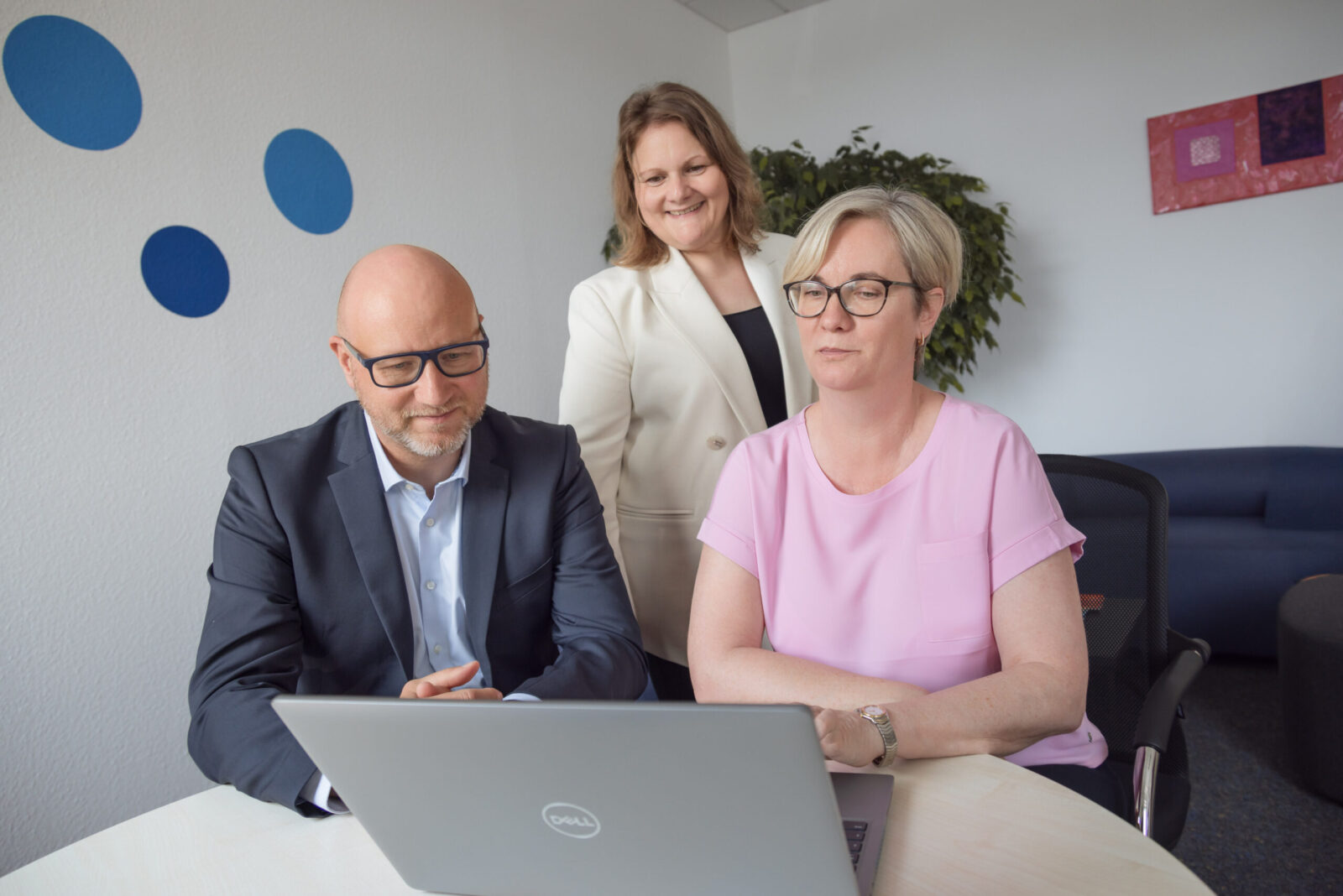Drei Personen im Gespräch an einem Besprechungstisch, fokussiert auf einen Laptop – professionelle Zusammenarbeit in angenehmer Büroumgebung.