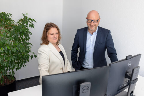 Zwei Personen in Businesskleidung stehen gemeinsam an einem Schreibtisch und betrachten konzentriert einen Bildschirm – modernes Büro mit freundlicher Atmosphäre.